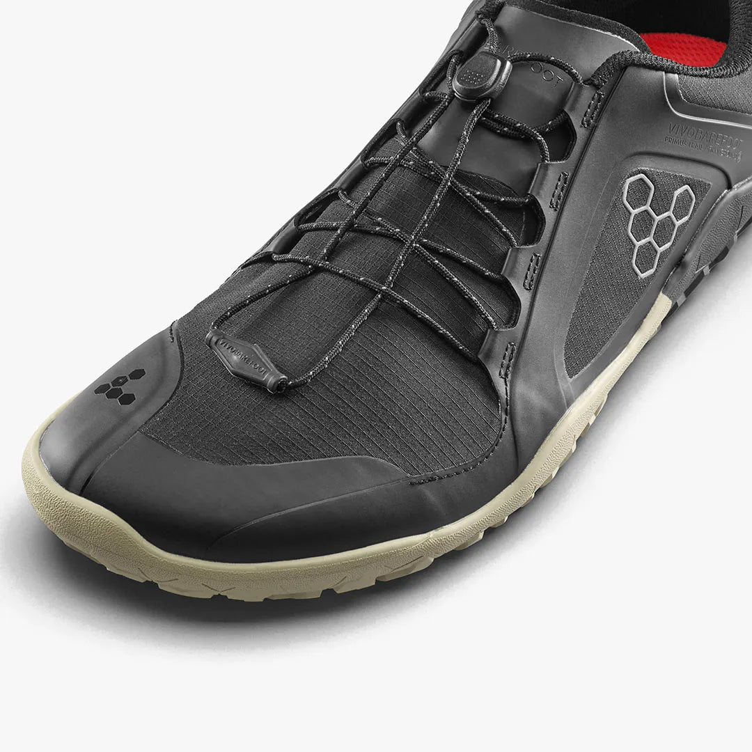 Close-up of the VIVOBAREFOOT Primus Trail III All Weather FG (Women's) in black with a beige sole, showcasing a hexagonal logo and elastic lace system—water-resistant sneakers crafted for trail running.