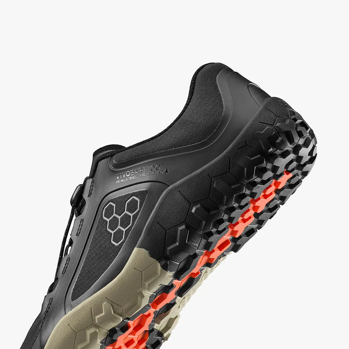 Close-up of the VIVOBAREFOOT Primus Trail III All Weather FG (Women's), a black and beige athletic shoe featuring a hexagonal tread pattern and orange accents on the sole. These water-resistant sneakers are designed for enhanced performance and are displayed against a white background.