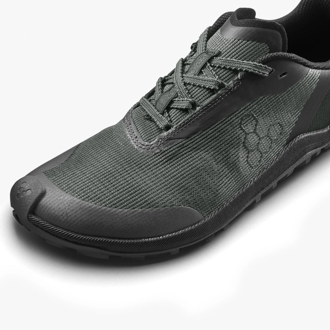 A close-up of the VIVOBAREFOOT Primus Trail Flow (Women's), a dark green minimalist athletic shoe featuring textured fabric, an all-terrain black outsole, hexagon side pattern, and neatly tied laces.