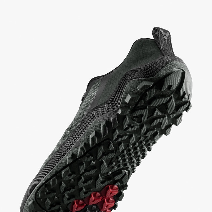 A close-up of the VIVOBAREFOOT Primus Trail Flow (Women's) in black, angled upward to showcase its lightweight design and rugged, multidirectional outsole with a red section for improved grip.