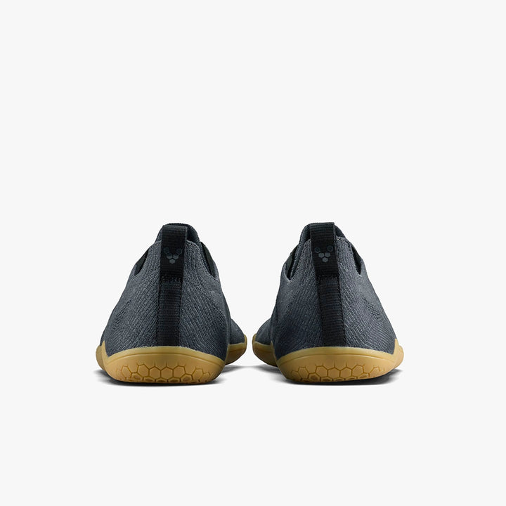 The Primus Lite Knit Natural (Women's) by STRIDE LAB USA is a pair of dark gray barefoot performance trainers with tan rubber soles, textured fabric, recycled PU insoles, and heel pull tabs, shown from the back on a white background.