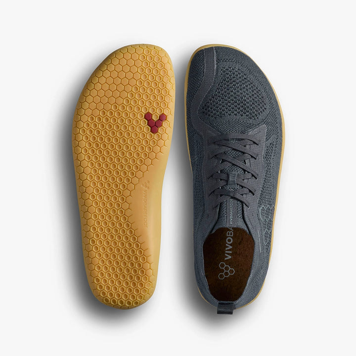 The STRIDE LAB USA Primus Lite Knit Natural (Women's) trainers feature dark gray mesh-like uppers with laces for style and flexibility, while their yellow soles display a honeycomb pattern and small red logo.