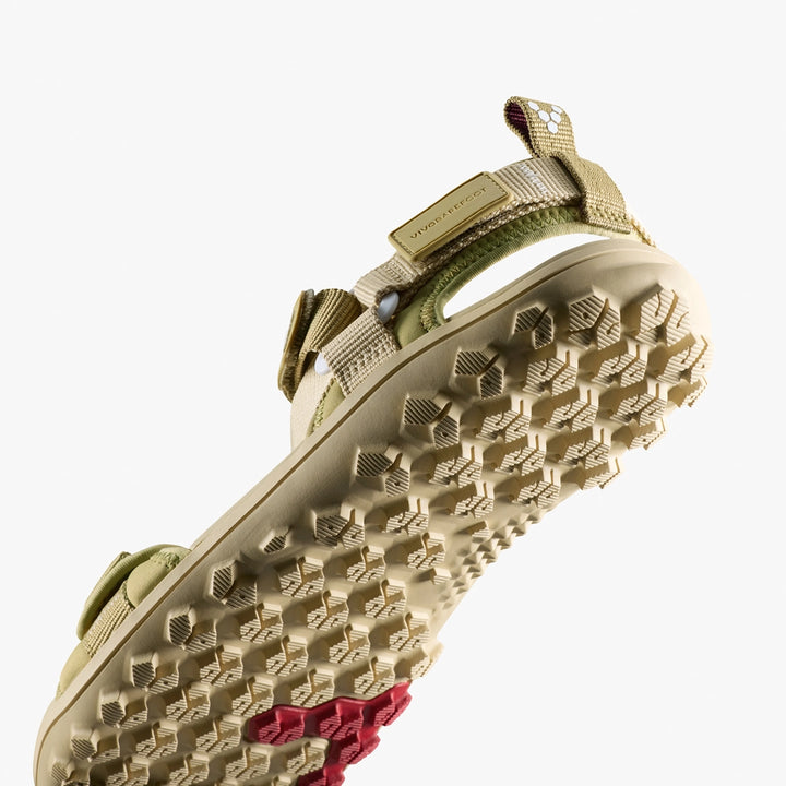 A close-up of the VIVOBAREFOOT Tracker Ora Sandal (Women's) shows beige straps and a rugged sole with hexagonal patterns and a red accent, photographed from below against a white background.