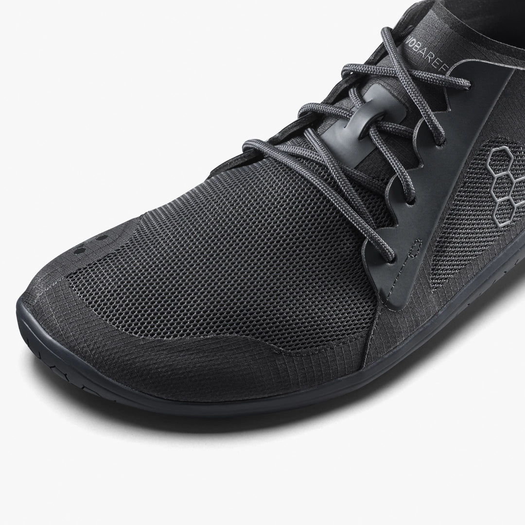 A close-up of the VIVOBAREFOOT Primus Lite IV (Men’s), a black performance trainer with a mesh upper, black laces, textured sole, subtle design details, recycled materials, and a small hexagonal logo on the side.