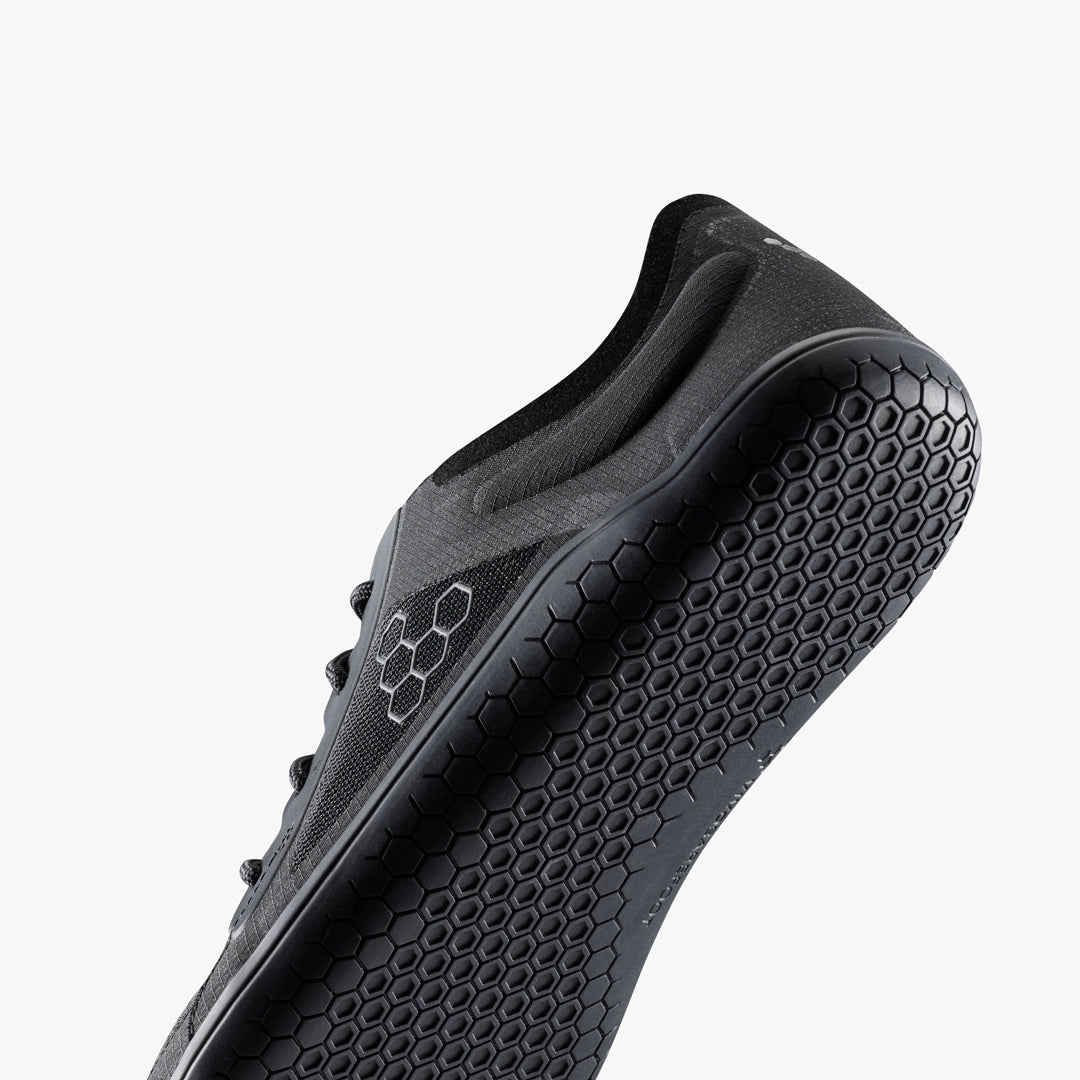 A close-up of the VIVOBAREFOOT Primus Lite IV (Men's) trainer from below shows its textured honeycomb-patterned sole, hexagonal side logo, and recycled materials, all set against a plain white background.