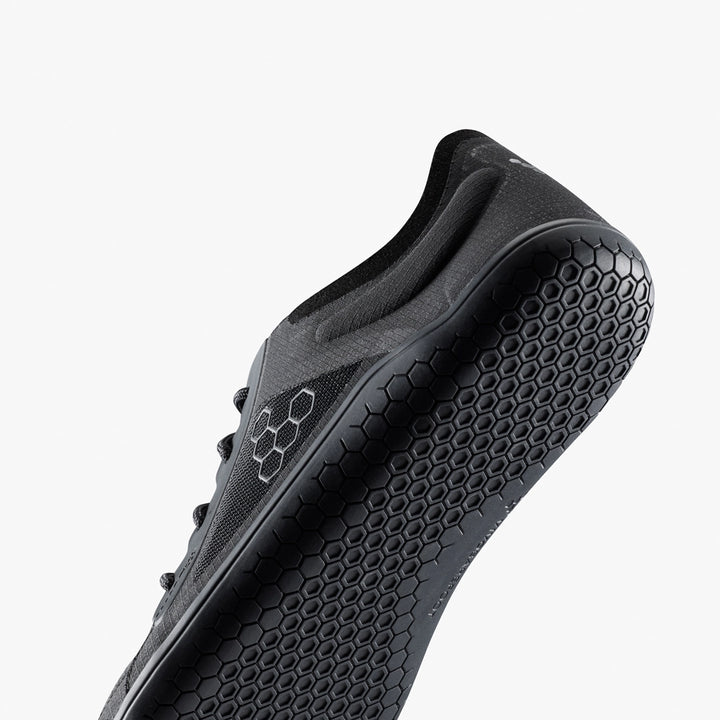 A close-up of the VIVOBAREFOOT Primus Lite IV (Men's) trainer from below shows its textured honeycomb-patterned sole, hexagonal side logo, and recycled materials, all set against a plain white background.