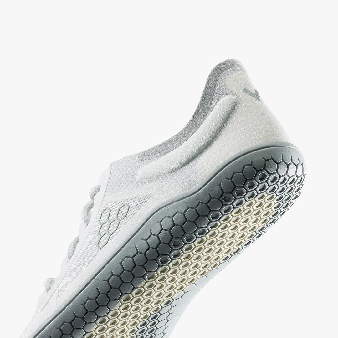 A low-angle close-up of the VIVOBAREFOOT Primus Lite IV (Men's), a white athletic shoe with a gray honeycomb-patterned sole, crafted from recycled materials, features subtle branding and minimalist design against a white background.