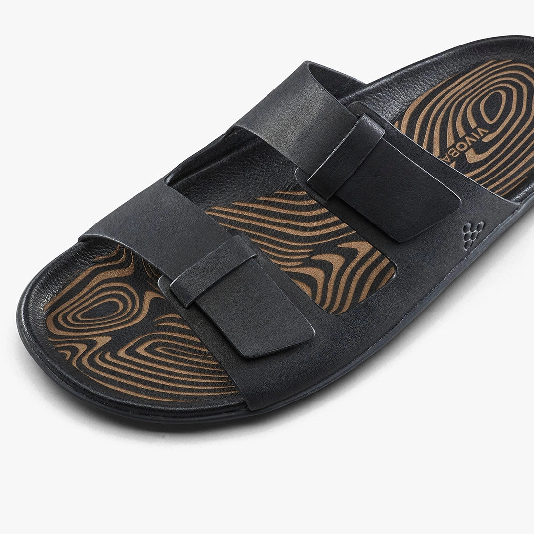 The VIVOBAREFOOT Sensus Yin Slide (Men's) in black features two adjustable straps and a patterned brown and black insole made from chrome-free Taro leather, displayed on a plain white background.