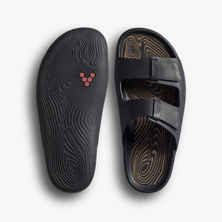 A pair of black VIVOBAREFOOT Sensus Yin Slide (Men's) sandals with dual straps. One sandal is flipped to reveal a textured sole and small pink logo; the other shows a top view with woodgrain-style insole detailing.