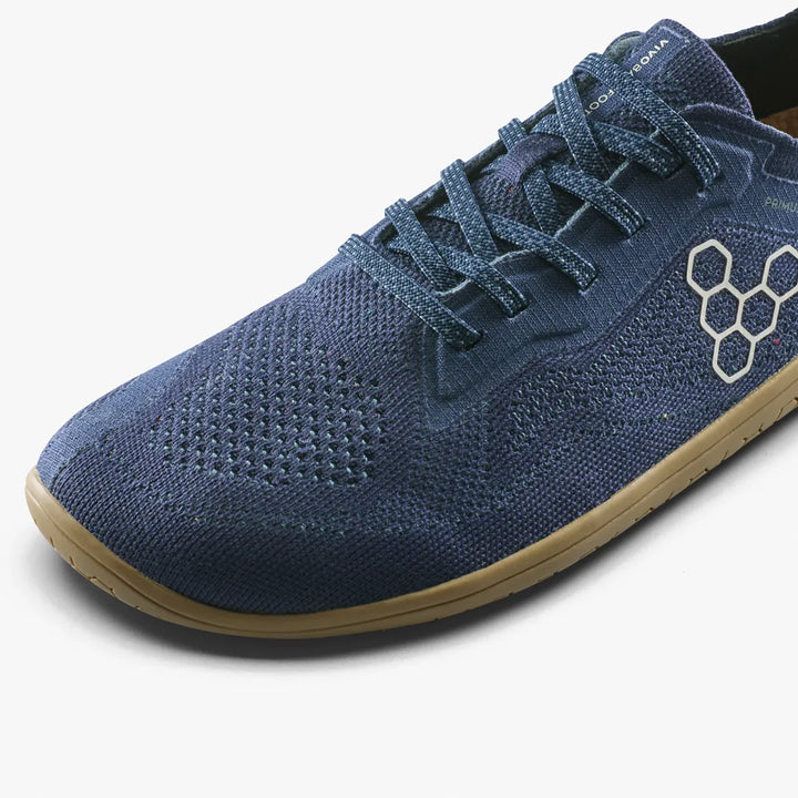 A close-up view of the VIVOBAREFOOT Primus Lite Knit Natural (Men’s) barefoot performance trainer in blue, featuring a textured knit upper, hexagonal logo detail, blue laces, and a light brown sole on a white background.