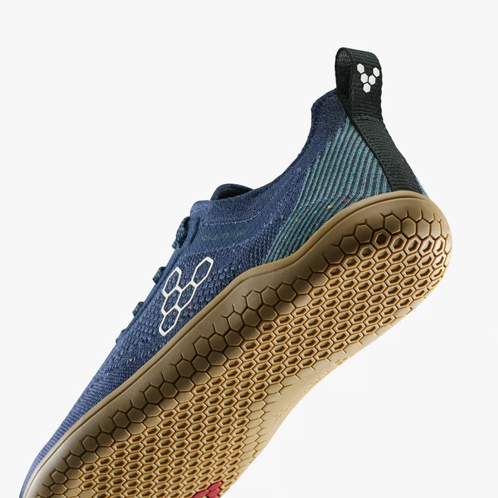 The VIVOBAREFOOT Primus Lite Knit Natural (Men’s) barefoot performance trainer in blue is displayed from a low angle, emphasizing its hexagonal-patterned tan sole. The heel tab features a white hexagon logo set against a black background.