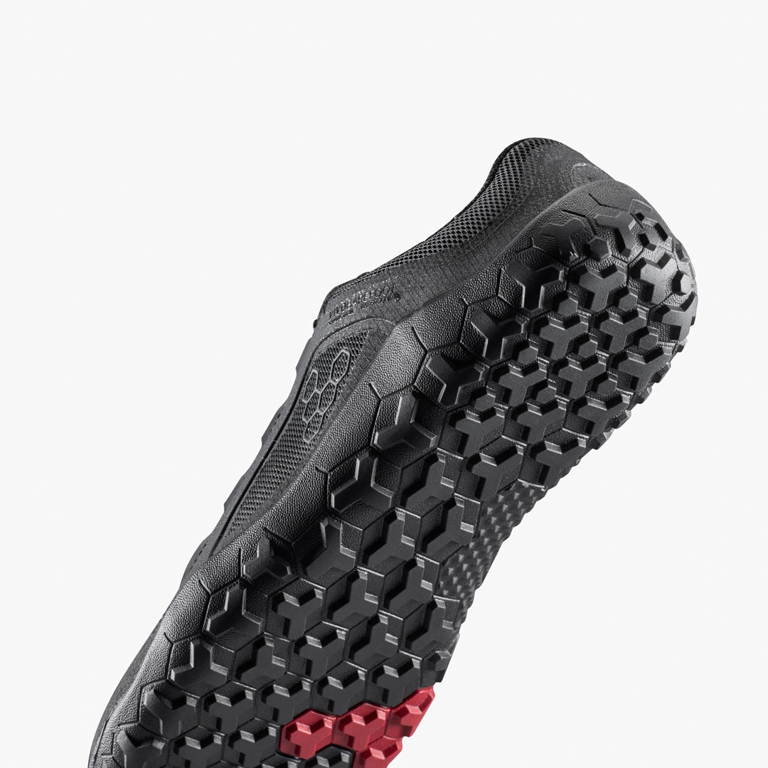 A close-up of the Primus Trail FG 3.5 (Men's) by VIVOBAREFOOT shows its black sole with a textured hexagonal tread, a small red hexagon detail, and recycled material construction, set against a plain white background.