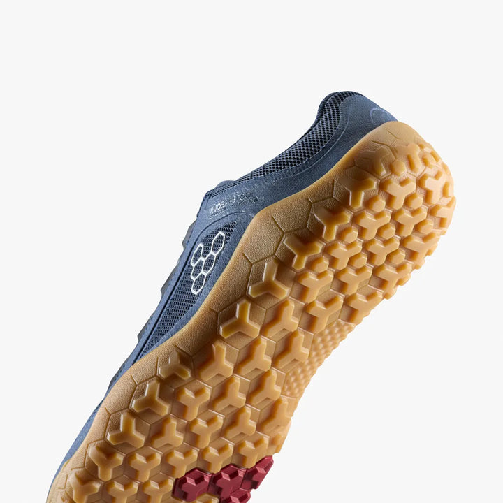 The VIVOBAREFOOT Primus Trail FG 3.5 (Men’s) is shown from below in blue, highlighting its tan, hexagonal-patterned firm ground outsole with a small red accent near the arch, set against a plain white background.