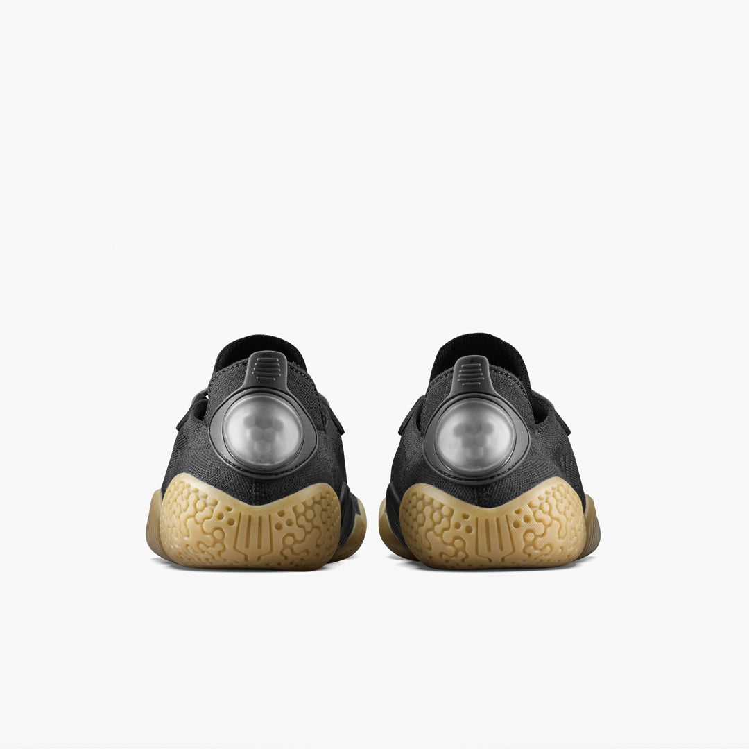 The VIVOBAREFOOT Motus Flex Natural (Men's) calisthenics shoes in black feature tan natural rubber outsoles for added traction and large translucent circles on the heels. Shown from the rear against a plain white background.