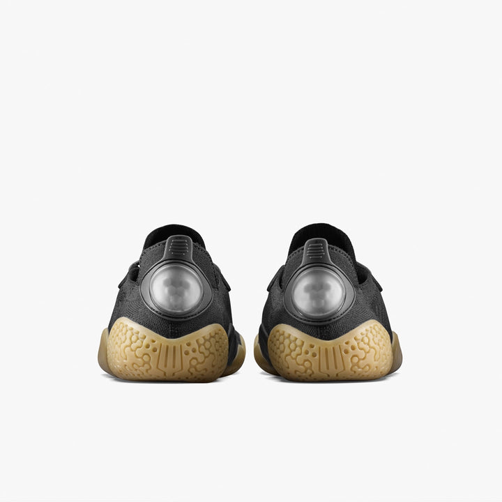 The VIVOBAREFOOT Motus Flex Natural (Men's) calisthenics shoes in black feature tan natural rubber outsoles for added traction and large translucent circles on the heels. Shown from the rear against a plain white background.
