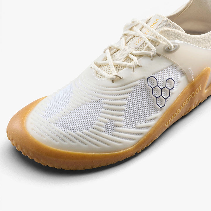 The VIVOBAREFOOT Motus Strength II (Men's) is a white barefoot trainer with a tan sole, hexagonal logo, and VIVOBAREFOOT branding, designed for grip and stability during high-impact workouts, shown from a top-side angle on white.
