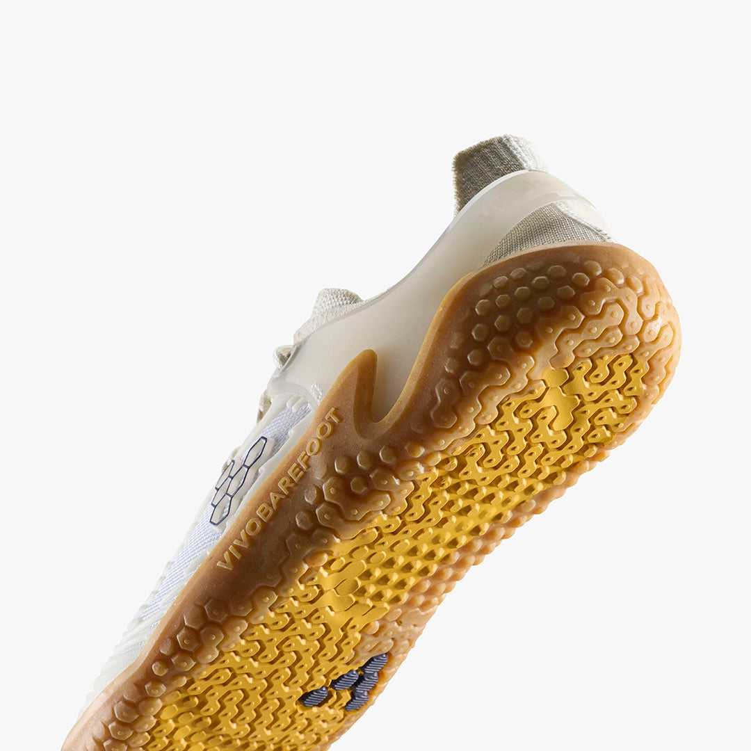 A close-up of the Motus Strength II men's trainer from VIVOBAREFOOT shows its white sole with textured, yellow-brown hexagonal grip patterns for superb traction and stability. A geometric design accents the side, with a plain white background.
