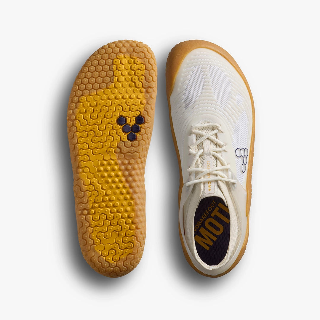 A pair of VIVOBAREFOOT Motus Strength II (Men's) barefoot trainers in white with yellow, textured soles. One shoe is shown from the top, the other from the bottom, highlighting a hexagonal logo for improved grip and stability.