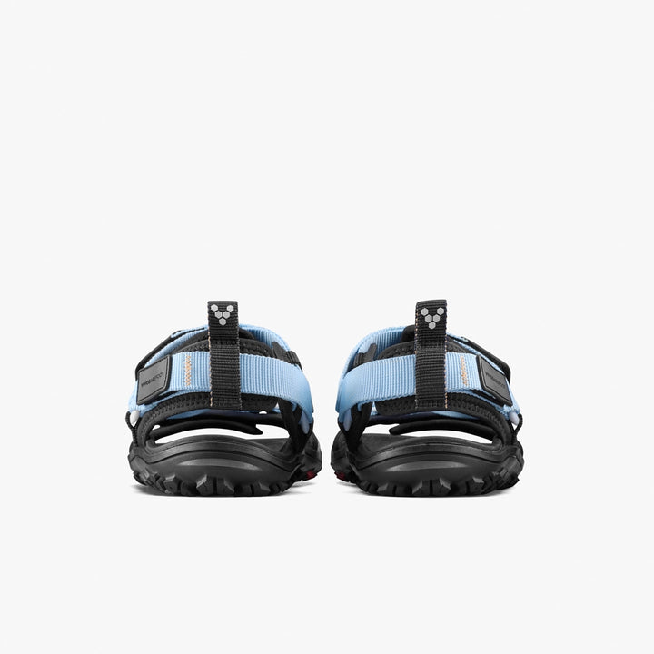 The VIVOBAREFOOT Tracker Ora Sandal (Men's) is an open-toe all-terrain sandal with light blue straps, rugged black soles, and a small logo on the heel strap, shown here from the back.