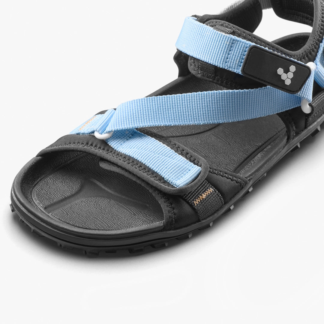 A close-up of the VIVOBAREFOOT Tracker Ora Sandal (Men's), featuring a black barefoot design, all-terrain textured outsole, and light blue adjustable straps with a small black and white side logo.