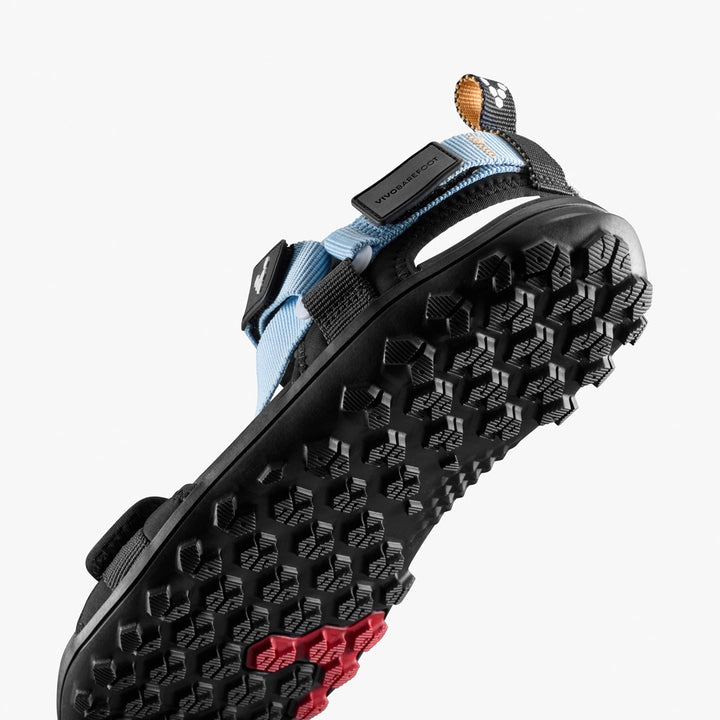 A close-up shows the sole of the VIVOBAREFOOT Tracker Ora Sandal (Men's) with rugged, textured black tread, blue and black straps, and a small red heel section against a white background.