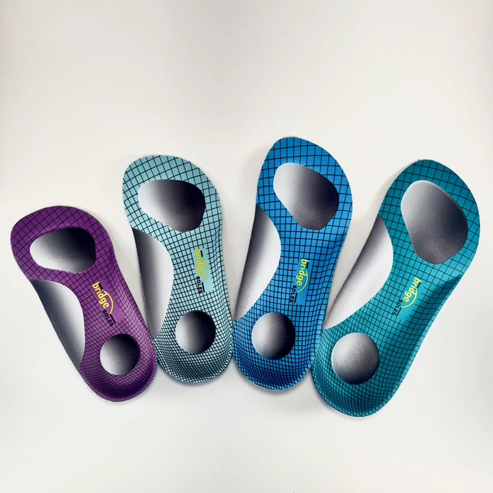 Four vibrant shoe insoles from Bridge Soles, two in purple and two in blue, are arranged side by side on a white background. Each Bridge Soles insole features a grid pattern and highlighted foot support zones, with the brand name “Bridge Soles” clearly visible.