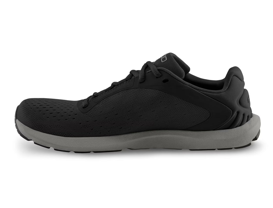 The TOPO ST-6 (Men's) athletic shoe is shown in side profile on a white background, featuring a black perforated upper, black laces, thick gray sole, and a roomy toe box.