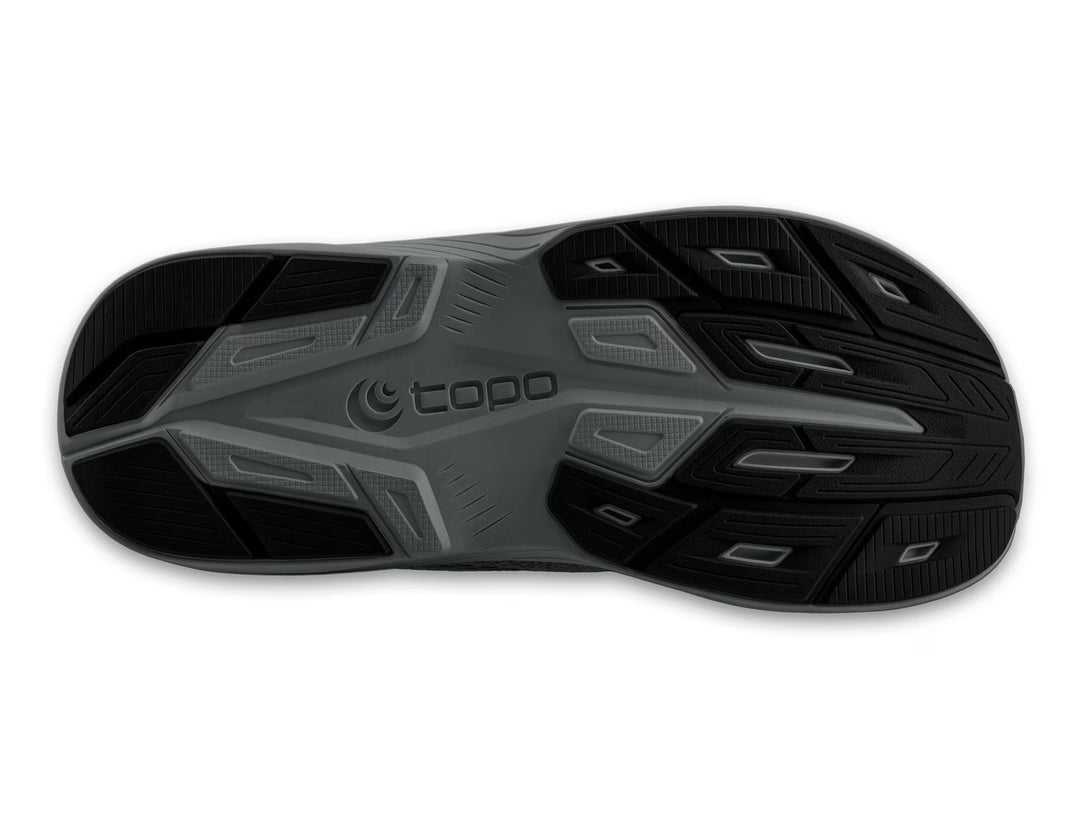 The image shows the black and gray rubber outsole of the TOPO Magnifly 6 (Men's) running shoe, with textured tread patterns and the TOPO brand name in raised letters at the center.