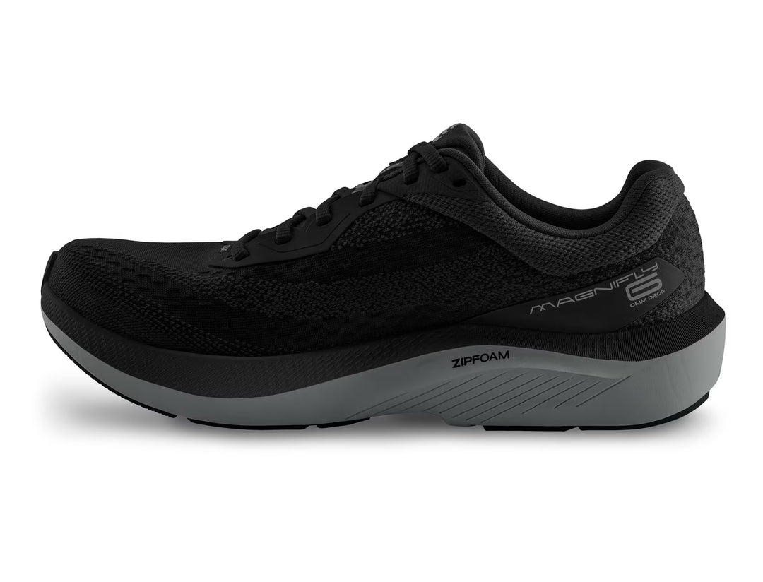 The TOPO Magnifly 6 (Men's) is a black cushioned running shoe featuring a textured mesh upper, black laces, and a thick two-tone gray and black sole. "MAGNIFLY 6" is on the side, with a responsive ZipFoam midsole for added comfort.