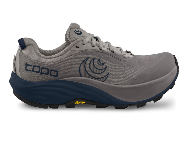 The TOPO Pursuit 3 (Men’s) is a gray and navy trail runner with “topo” and a circular logo on the side, designed with a Vibram Megagrip sole for enhanced traction on rugged terrain.