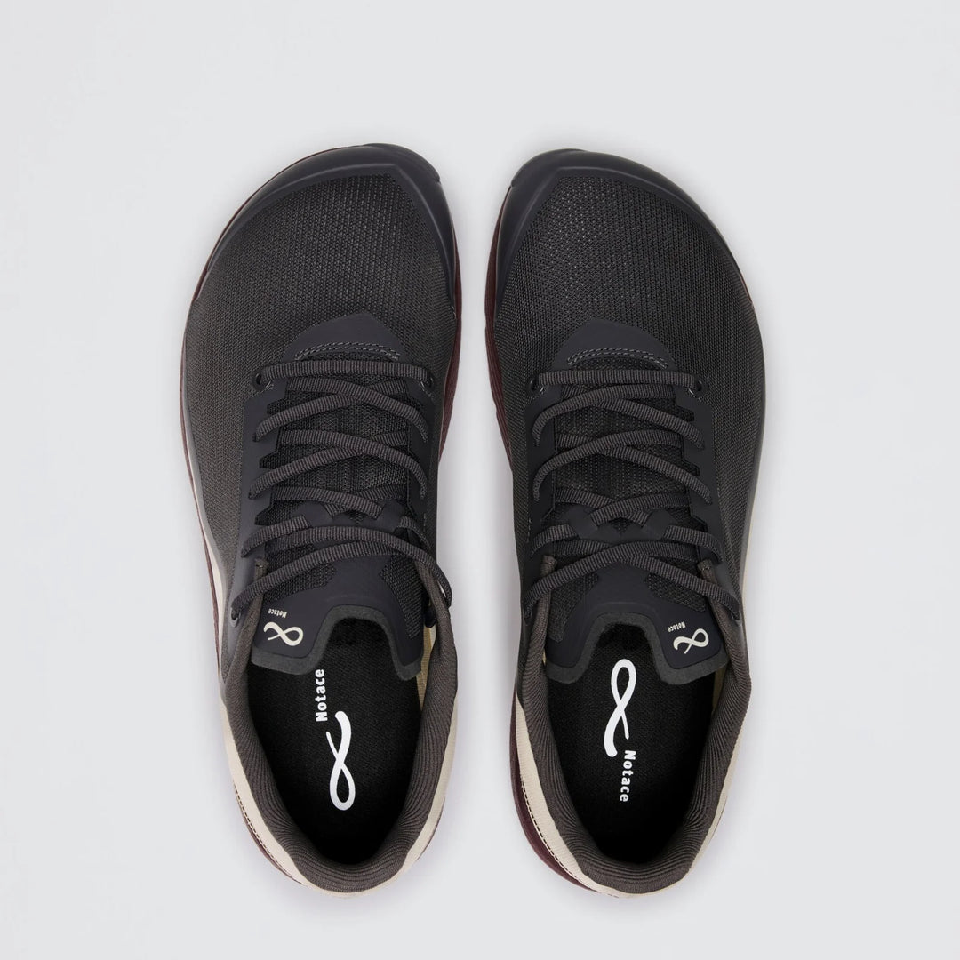 A pair of Notace Michi 1 (Men’s) black athletic shoes with white soles and black laces, viewed from above on a plain light gray background. Designed as a flexible training shoe, they feature an eTPU midsole for enhanced comfort and responsiveness. The insoles display a white Notace logo and the word “Balance.”.