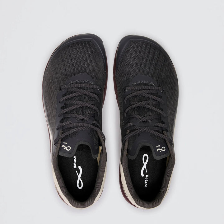 A pair of Notace Michi 1 (Men’s) black athletic shoes with white soles and black laces, viewed from above on a plain light gray background. Designed as a flexible training shoe, they feature an eTPU midsole for enhanced comfort and responsiveness. The insoles display a white Notace logo and the word “Balance.”.