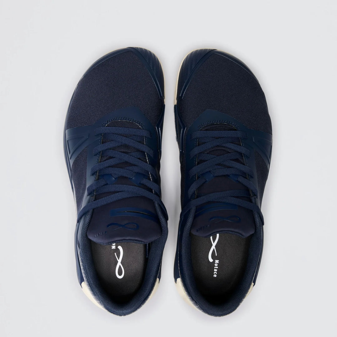 A pair of Notace Koto 1 (Women’s) dark blue athletic gym shoes with laces, viewed from above on a light gray background. The insoles feature a white infinity symbol and the word “balance,” highlighting their multidirectional control ideal for agile movement.