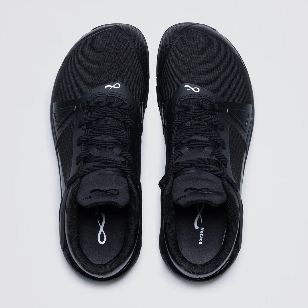 The Notace Koto 1 (Women's) is a pair of black athletic court shoes with black laces, white infinity logos on the tongue and insole, and a sleek, minimal design for multidirectional control and grounded stability on a light background.