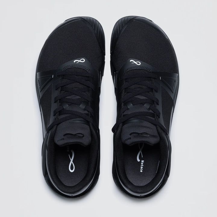 The Notace Koto 1 (Women's) is a pair of black athletic court shoes with black laces, white infinity logos on the tongue and insole, and a sleek, minimal design for multidirectional control and grounded stability on a light background.