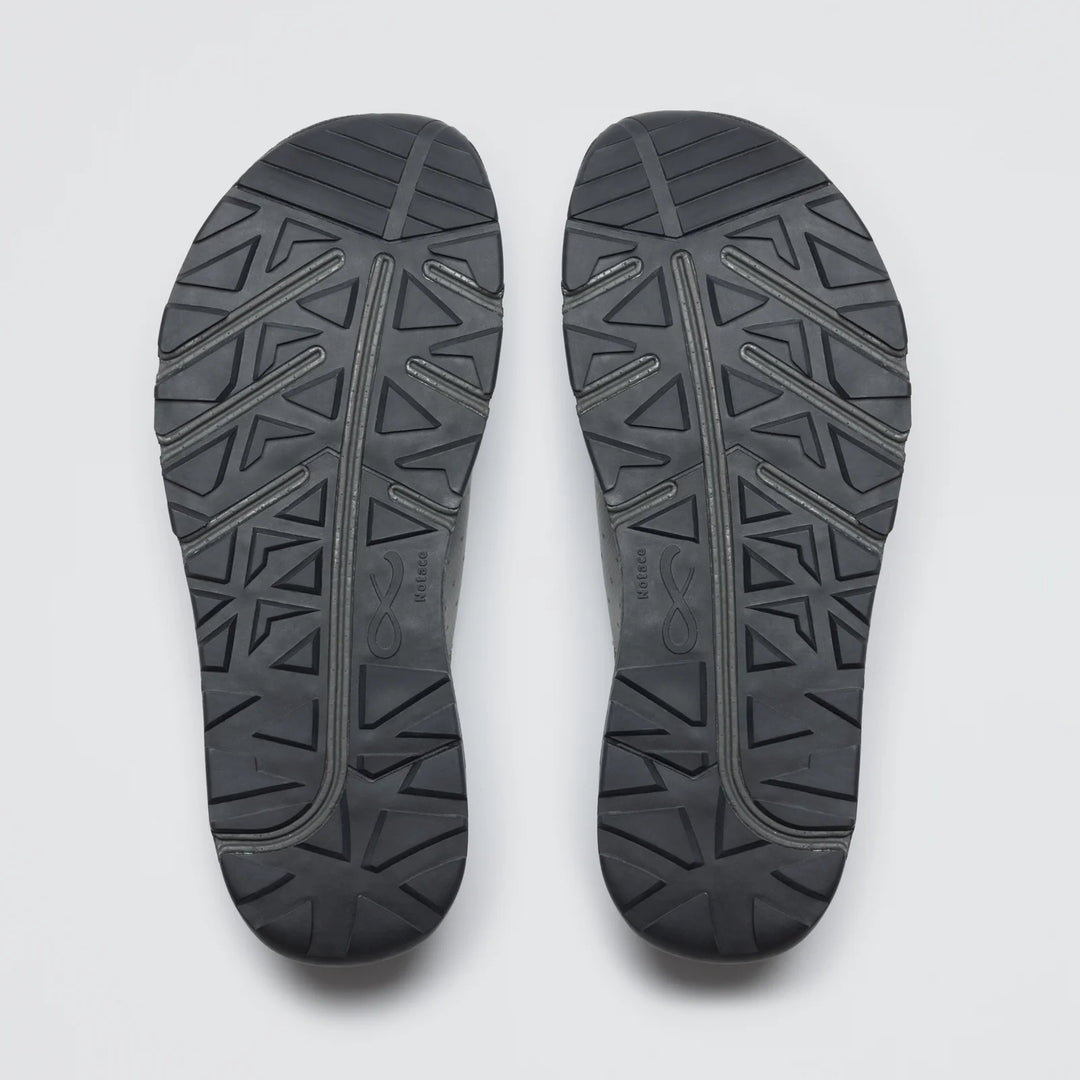 A pair of Notace Yama T1 men's trail shoe soles in black, featuring a geometric tread with triangles and diagonal lines, designed for all-day comfort, are displayed side by side on a plain white background.