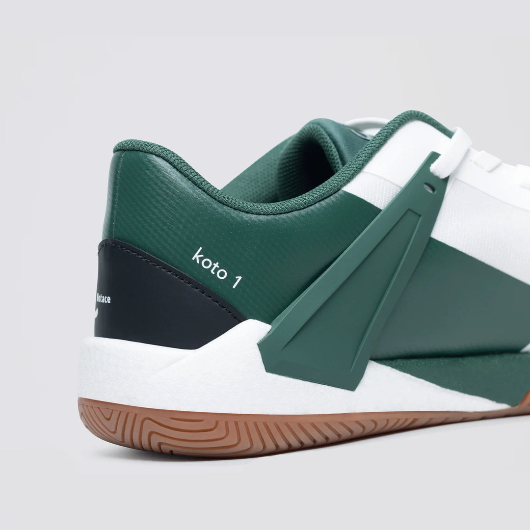 Close-up of the heel of the Notace KOTO 1 (Men's) court shoe in green, white, and black with "koto 1" text on the side. Features a geometric design, white sole, brown textured outsole, and multidirectional control.