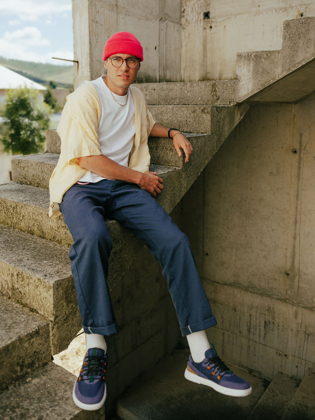 A man in Barebarics Revive (Men's) lightweight shoes, a red beanie, glasses, white t-shirt, yellow overshirt, and blue pants sits on concrete steps in an unfinished building, looking at the camera.
