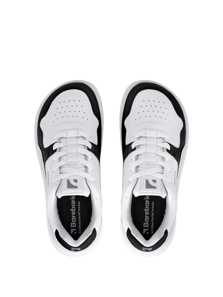 A pair of white Barebarics Zing (Unisex) sneakers with black accents, seen from above on a light gray background. They feature perforated toe boxes, white laces, and Barebarics branding on the insoles for barefoot quality.