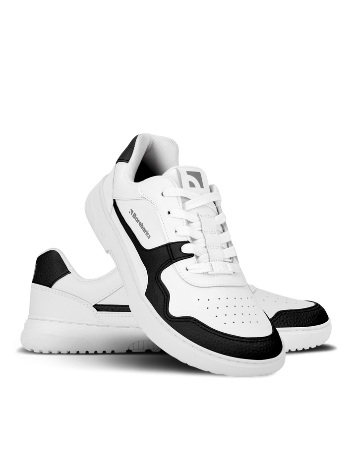 The Barebarics Zing (Unisex) sneakers feature a white design with black accents on the toe, heel, and sides. One shoe stands upright as the other is tilted to showcase its sole and perforated sustainable leather detailing.