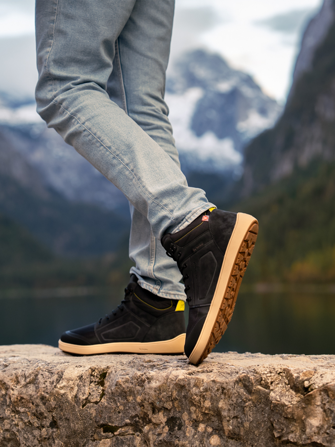Wearing BeLenka's ArcticEdge (Unisex) waterproof barefoot shoes and light blue jeans, a person stands on a stone ledge with one leg crossed, surrounded by scenic mountains, a lake, and trees.