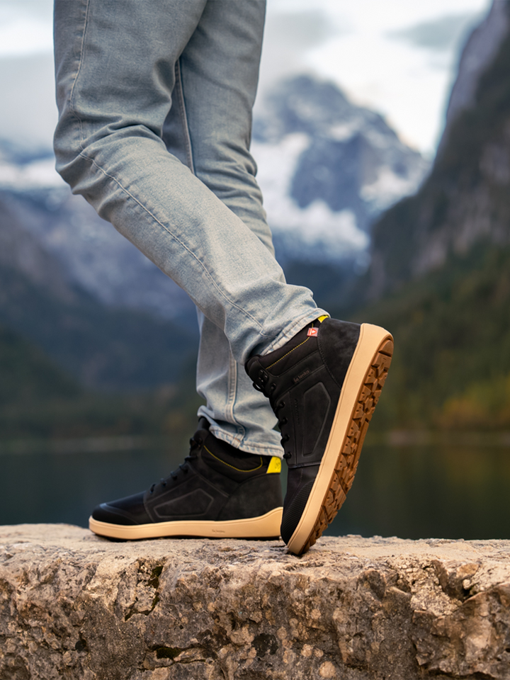 Wearing BeLenka's ArcticEdge (Unisex) waterproof barefoot shoes and light blue jeans, a person stands on a stone ledge with one leg crossed, surrounded by scenic mountains, a lake, and trees.