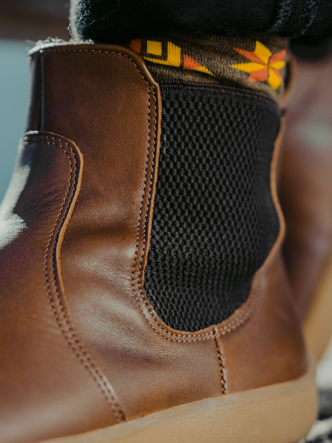 Close-up of the BeLenka Entice Neo (Unisex) boot in brown leather, featuring a black elastic panel. Styled with black pants and vibrant socks showcasing a geometric yellow and red pattern, these barefoot boots offer both comfort and style with their wide toe box and zero-drop sole.
