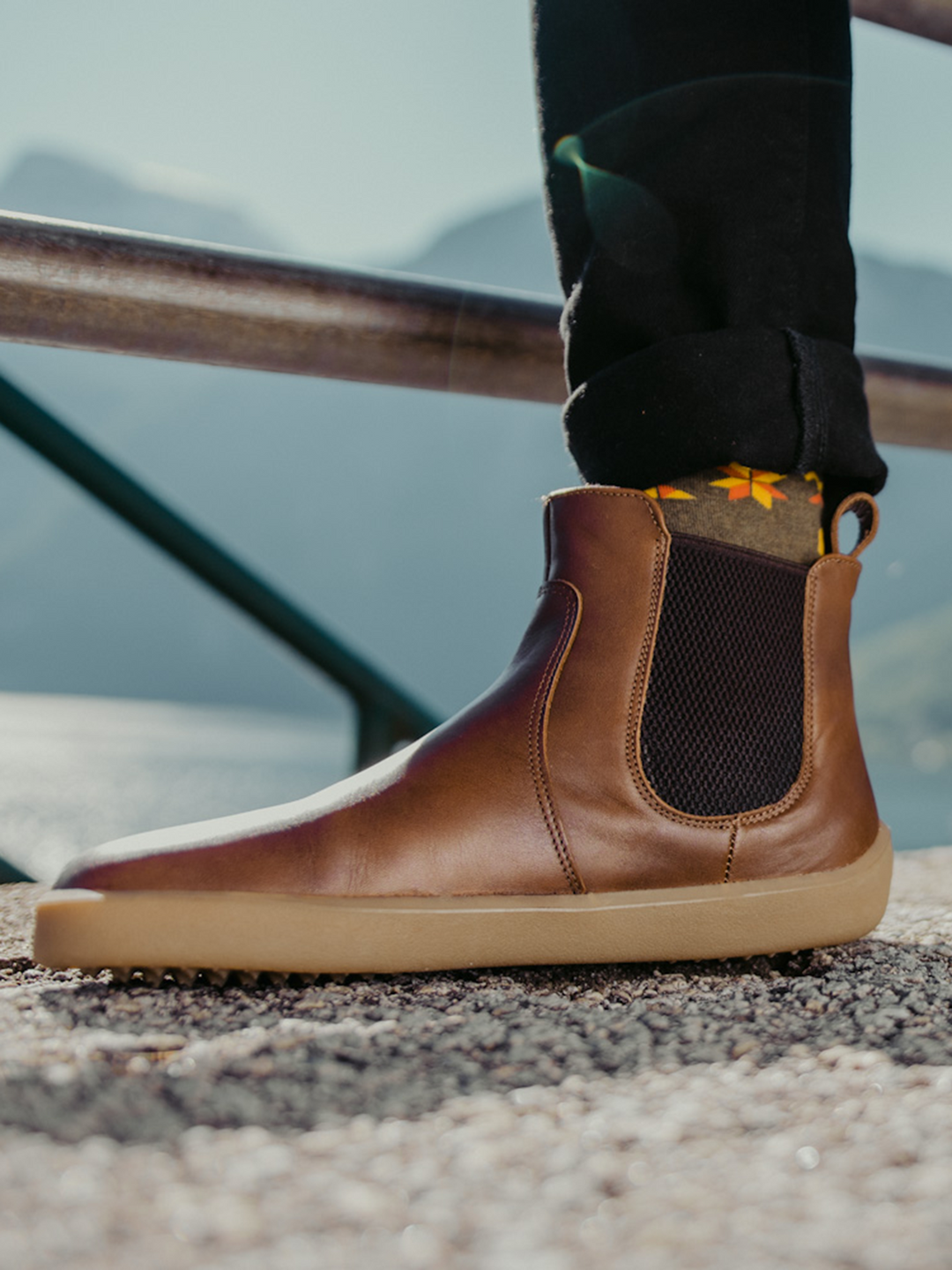 A close-up of a person wearing BeLenka Entice Neo (Unisex) brown leather Chelsea boots with a wide toe box, paired with black pants and yellow patterned socks, standing outdoors on a gravel surface with mountains and water in the background.