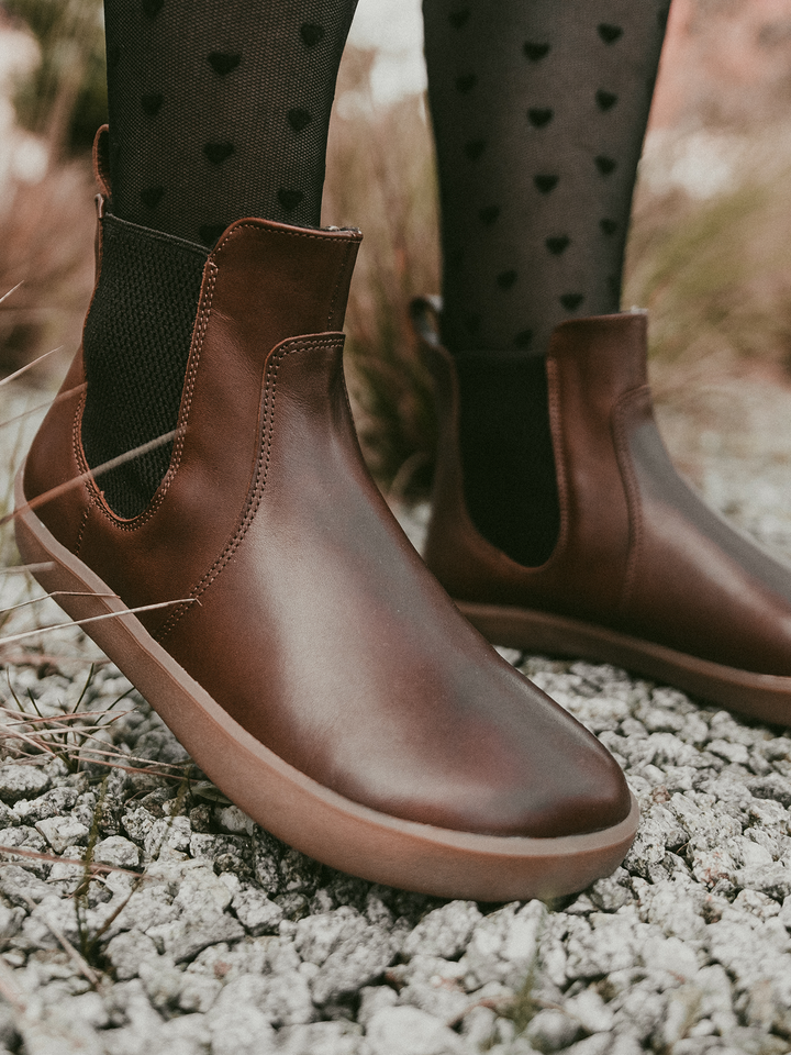 Close-up of a person standing outdoors on gravel among grass, wearing BeLenka Entice Neo (Unisex) brown leather ankle boots with a wide toe box and black heart-patterned tights.