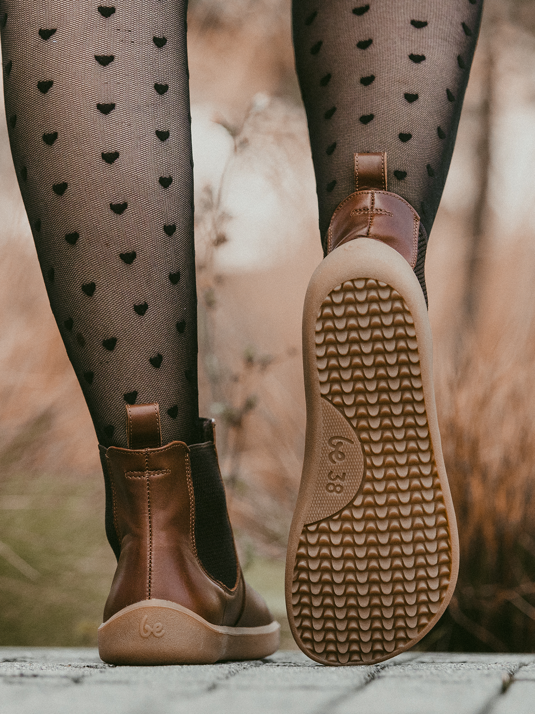 Close-up of a person wearing sheer black tights with small heart patterns and BeLenka Entice Neo (Unisex) brown barefoot boots featuring a zero-drop sole, shown from behind while walking outdoors.