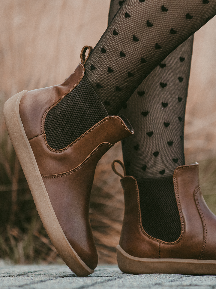 BeLenka’s Entice Neo (Unisex) brown ankle boots, designed with a wide toe box and tan soles, are paired with sheer black tights adorned with a heart pattern. The blurred background features outdoor foliage.