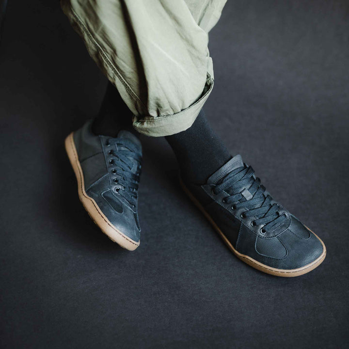 A person wearing light green pants, black socks, and a pair of BluSun BLSN-200 women’s barefoot shoes in dark navy with gum soles stands on a dark surface. The sneakers feature an organic cotton lining and a non-slip TPU outsole from BluSun, offering added comfort and stability.