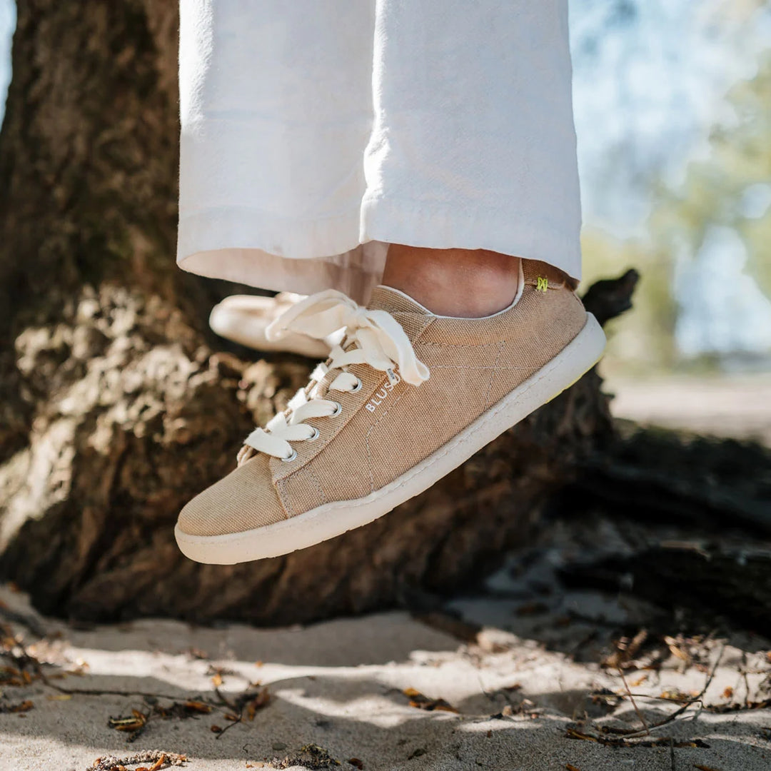 A person wearing BluSun BLSN-103 men’s beige vegan shoes with white laces and white pants stands on sand near a tree, with sunlight filtering through the trees in the background.