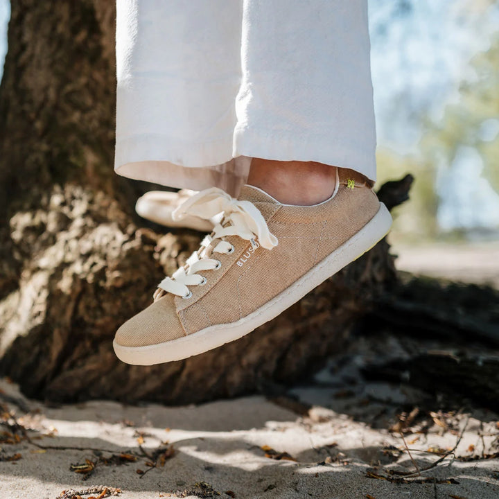 A person wearing BluSun BLSN-103 men’s beige vegan shoes with white laces and white pants stands on sand near a tree, with sunlight filtering through the trees in the background.